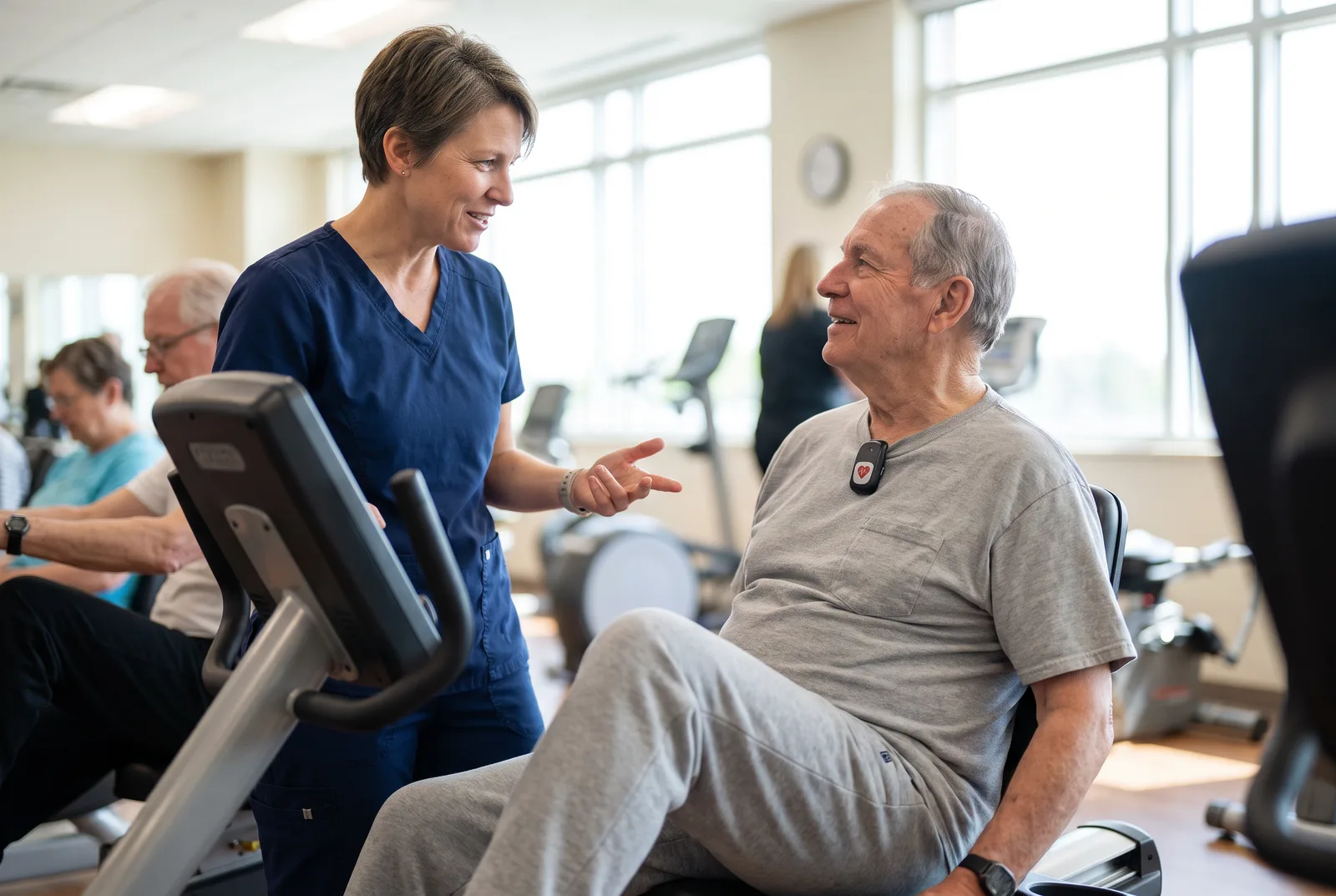 A clinical exercise physiologist in navy scrubs talks with an older man on a recumbent bike in a cardiac rehabilitation gym.