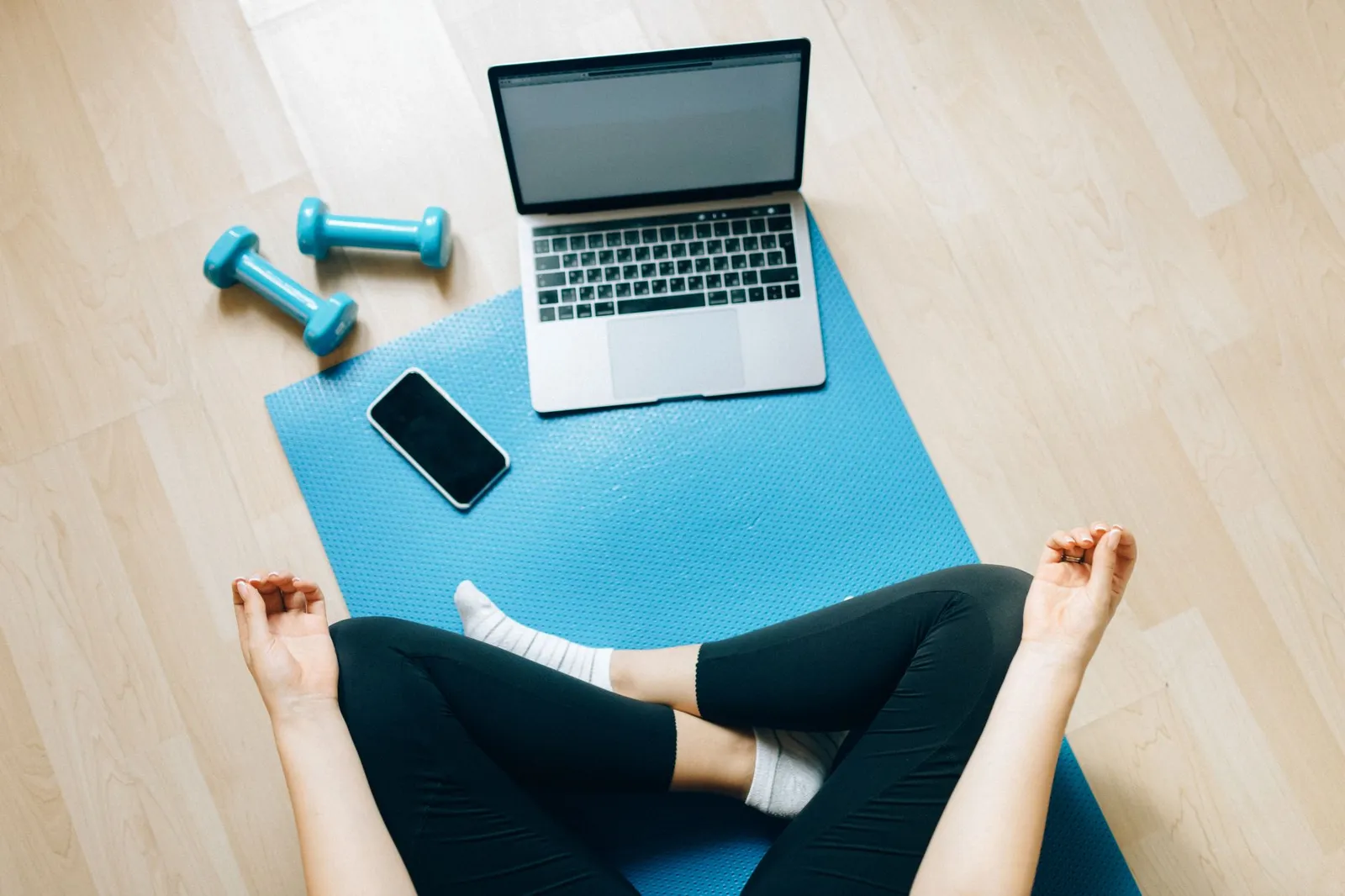 A person sits cross-legged on a blue yoga mat beside a laptop, a phone and two teal dumbbells on a wood floor.