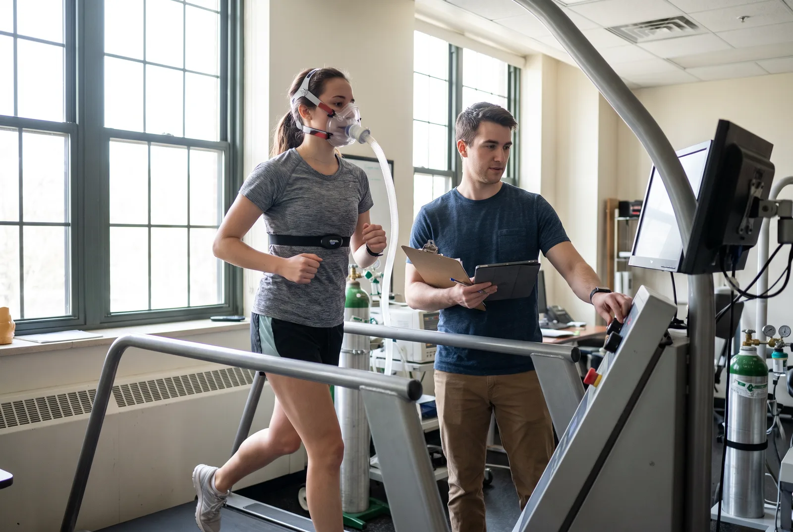 A student runs on a treadmill wearing a metabolic mask and heart rate strap while a graduate student with a clipboard adjusts the treadmill in a university exercise physiology lab.